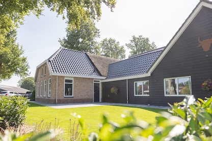 zijkant tuin Voorgevel van een modern landelijk hotel met een bakstenen gevel, donker puntdak en verzorgde groene tuin, omringd door hoge bomen op een zonnige dag.